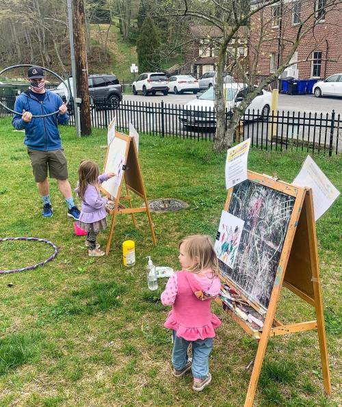 A man and two young children paint on easels in a grassy area, with cars and trees in the background.