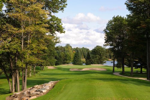 A scenic view of a golf course with lush green grass, sand traps, trees, and a lake under a partly cloudy sky.