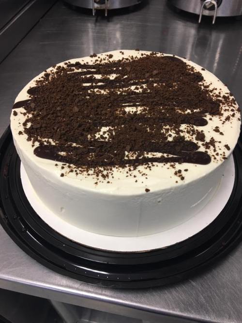 A round cake with white frosting, topped with chocolate drizzle and crushed cookies, on a black cake base.