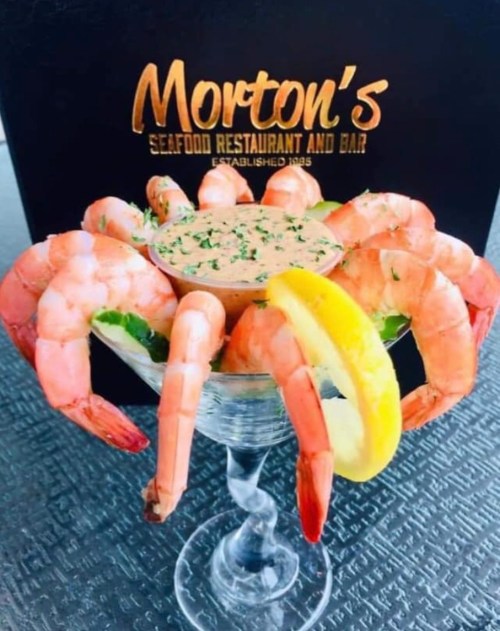 A martini glass filled with shrimp, garnished with lettuce, a lemon wedge, and a dipping sauce, against a restaurant backdrop.