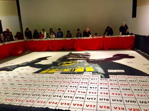 A large mosaic of a superhero made from colorful tiles, with people assembling it at a table in a spacious room.