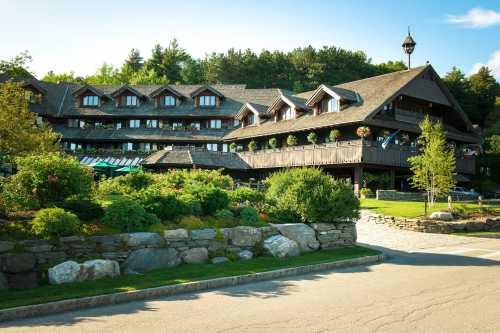 A charming lodge surrounded by greenery, featuring a wooden exterior and flower-filled balconies under a clear blue sky.