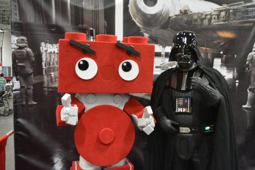 A red block character poses with a Darth Vader figure at a convention, with a space-themed backdrop.