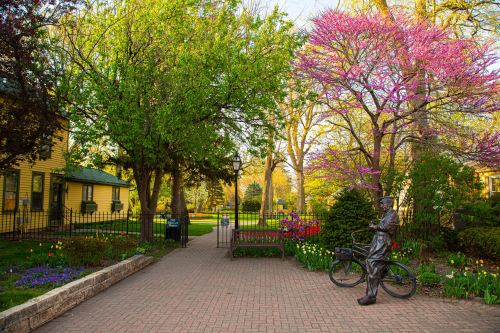 A peaceful park scene with vibrant trees, flowers, and a statue of a person with a bicycle along a brick pathway.
