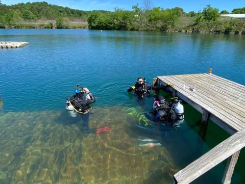 Two divers in wetsuits are submerged near a wooden dock in a clear blue lake, surrounded by greenery.
