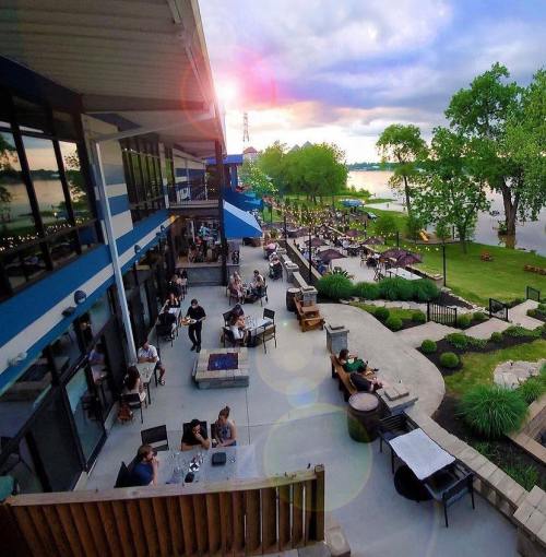 A lakeside venue with outdoor seating, people dining, and a sunset view over the water, surrounded by greenery.
