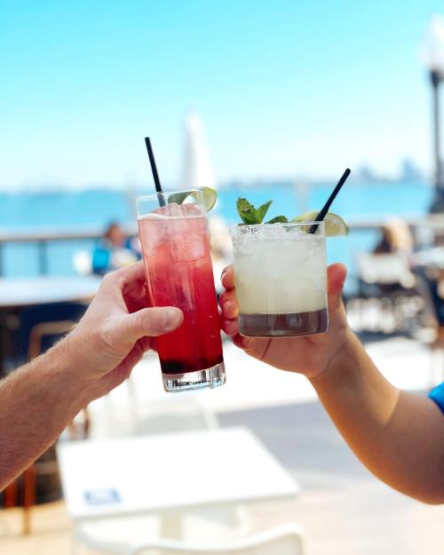 Two hands holding colorful cocktails with straws, against a bright blue sky and waterfront backdrop.