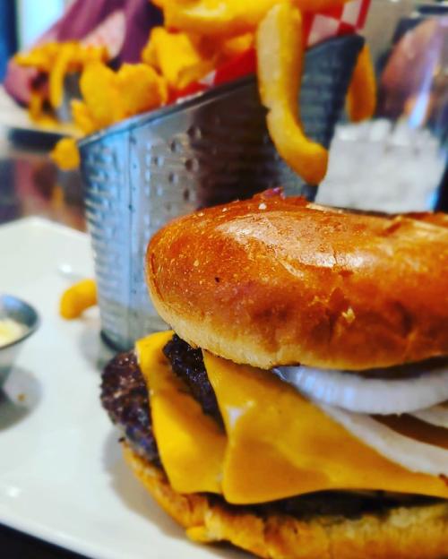 A close-up of a cheeseburger with melted cheese, onions, and a side of curly fries in a metal container.