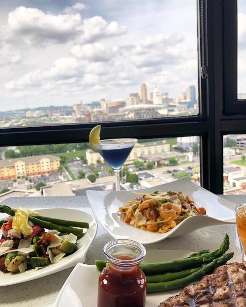 A table with gourmet dishes, including pasta, salad, and drinks, with a city skyline view in the background.