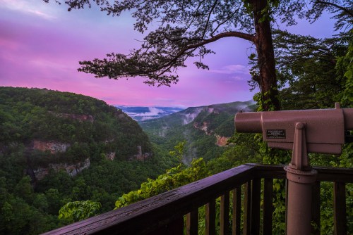 A scenic view of a lush valley at sunrise, with a telescope on a wooden deck overlooking the landscape.