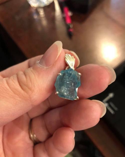 A hand holding a blue gemstone pendant with a silver setting, resting on a wooden surface.