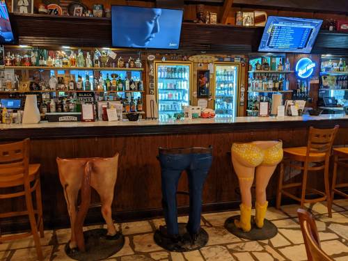 A bar with a wooden counter featuring quirky leg sculptures as stools, and a TV playing in the background.