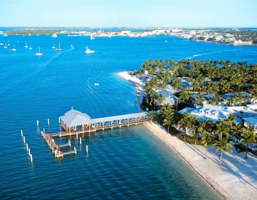 Aerial view of a coastal area with a pier, palm trees, and clear blue water, showcasing a serene beach landscape.