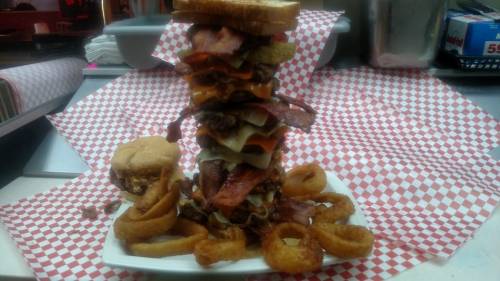 A towering burger stack with multiple patties, cheese, bacon, and onion rings on a plate, surrounded by checkered paper.