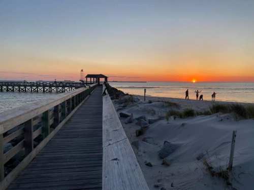 A wooden pier leads to a sunset over the water, with silhouettes of people enjoying the beach.