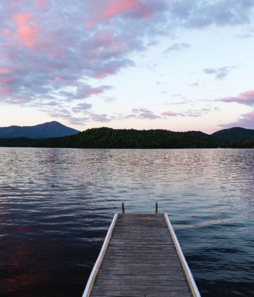 A wooden dock extends into a calm lake, surrounded by mountains and colorful clouds at sunset.