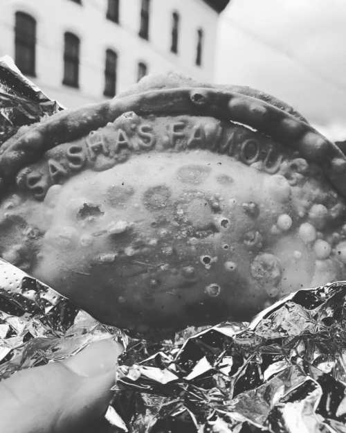 A hand holding a golden-brown pastry with "Sasha's Famous" embossed on it, wrapped in shiny foil.