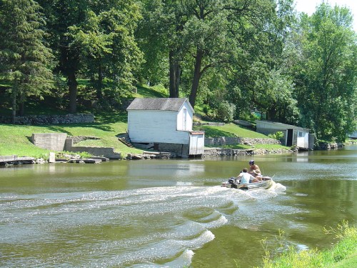 A small boat glides across a calm river, with green trees and a white shed in the background.