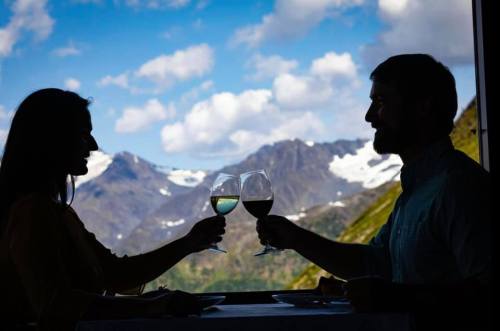 A couple toasting with wine glasses against a backdrop of mountains and a blue sky with clouds.