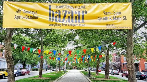 A banner for the Mainstrasse Village Bazaar, featuring colorful flags, dates, and location details.