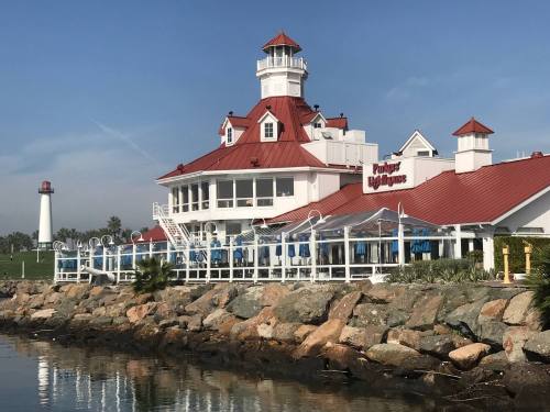 A waterfront building with a red roof and lighthouse nearby, surrounded by rocks and palm trees under a clear sky.