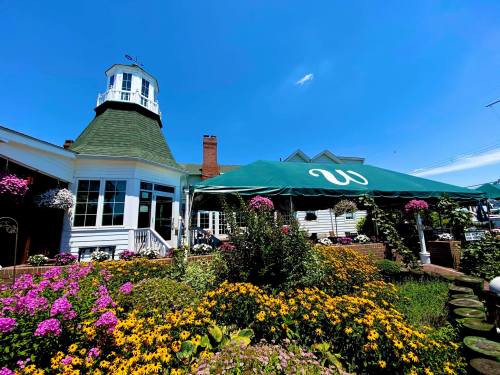 A charming restaurant with a green roof and outdoor seating, surrounded by colorful flowers under a clear blue sky.