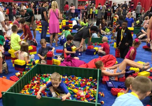 A vibrant scene of children and adults playing with colorful building blocks at a large event, surrounded by others.
