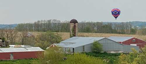 A colorful hot air balloon floats above a rural farm with red barns and a silos, surrounded by green fields and trees.