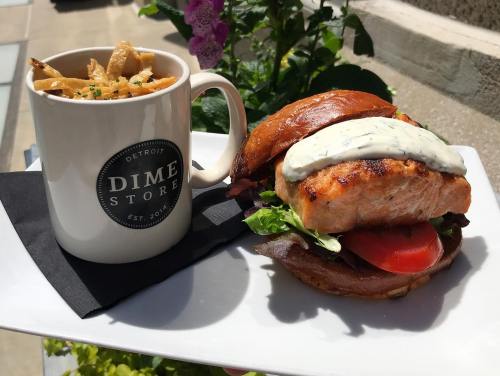 A salmon burger with lettuce and tomato on a plate, alongside a cup of fries and a mug from Dime Store.