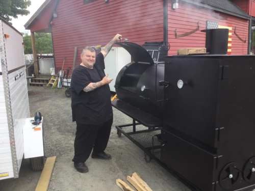 A man in a black shirt stands next to a large smoker, pointing at it with a smile, smoke rising from the top.