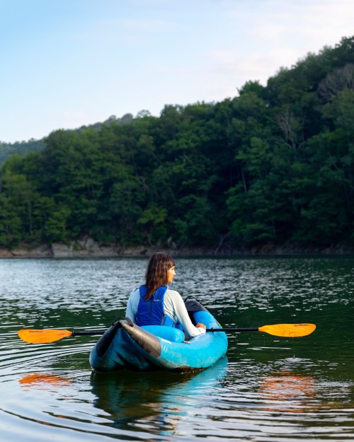 A person in a blue kayak paddles on a calm lake surrounded by green trees under a clear sky.