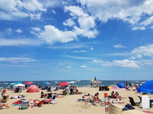 A busy beach scene with colorful umbrellas, sunbathers, and a clear blue sky with fluffy clouds.