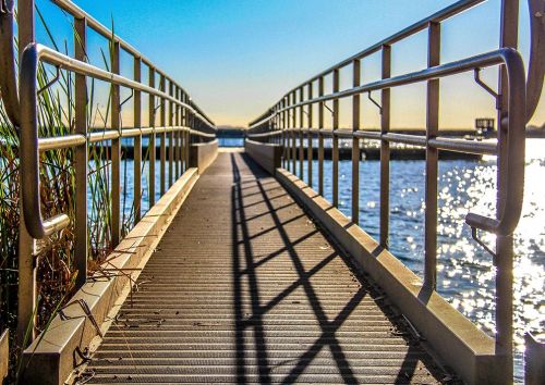 A sunlit pier extends over water, with metal railings and shadows creating a serene, inviting atmosphere.