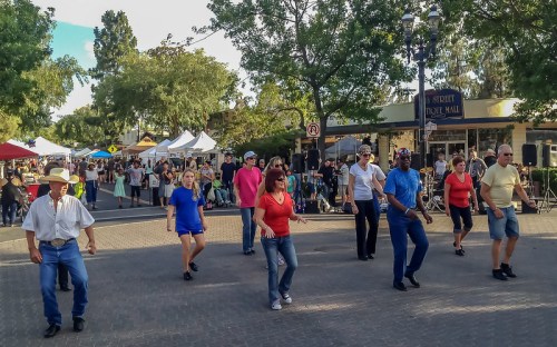A group of people dancing in a lively outdoor market, surrounded by trees and vendor tents.