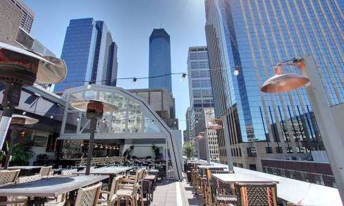 Rooftop dining area with tables, heaters, and a view of tall city skyscrapers under a clear blue sky.