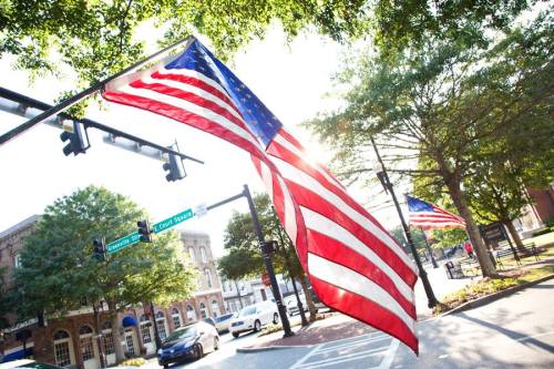 American flags wave in the breeze along a sunlit street with traffic lights and trees in the background.