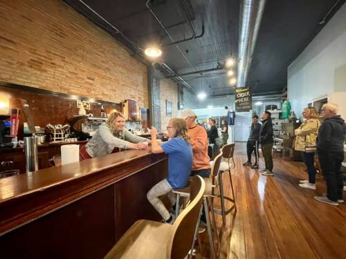 A barista serves customers at a coffee shop with a wooden counter and a line of people waiting.