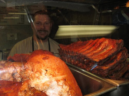 A smiling chef stands behind a glass display of delicious, smoked meats, including ribs and a large pork shoulder.
