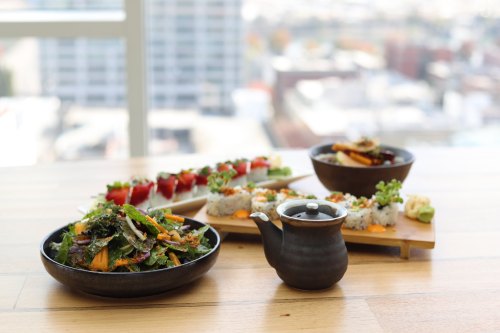 A table with a vibrant salad, sushi rolls, and a small teapot, set against a city skyline backdrop.