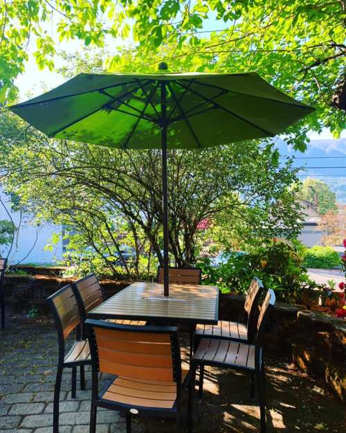 A shaded outdoor table with chairs under a green umbrella, surrounded by lush greenery and sunlight.