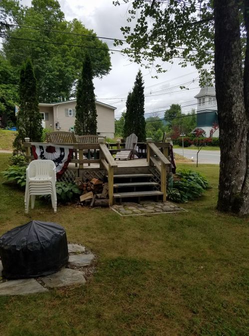 A wooden deck with chairs, surrounded by greenery and a fire pit, set in a residential area with trees and a cloudy sky.