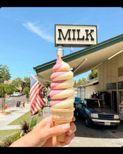 A hand holds a swirled pink and white ice cream cone in front of a shop with a "MILK" sign and an American flag.