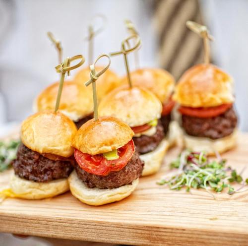 A wooden platter with mini burgers topped with tomatoes and pickles, garnished with fresh herbs.