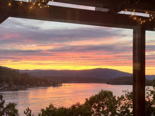 A vibrant sunset over a lake, framed by wooden beams and twinkling lights, with mountains in the background.