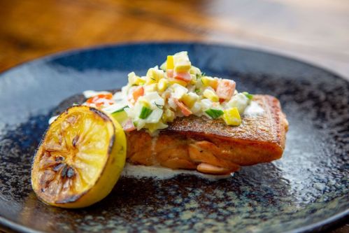 Grilled salmon fillet topped with fresh salsa and served with a charred lemon on a dark plate.