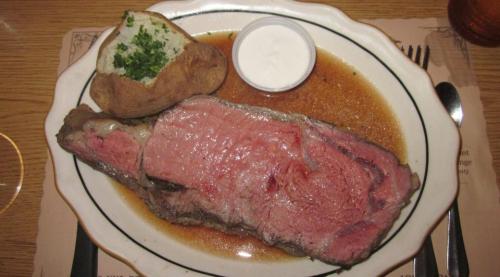 A plate with a large slice of prime rib, a baked potato with sour cream, and a side of gravy.