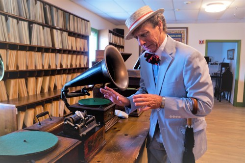 A man in a light suit and hat interacts with a vintage gramophone in a room filled with records.