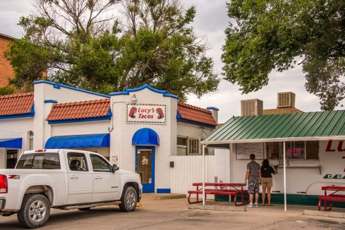 A white truck parked outside Lucy's Tacos, with two people ordering at a counter under a green awning.