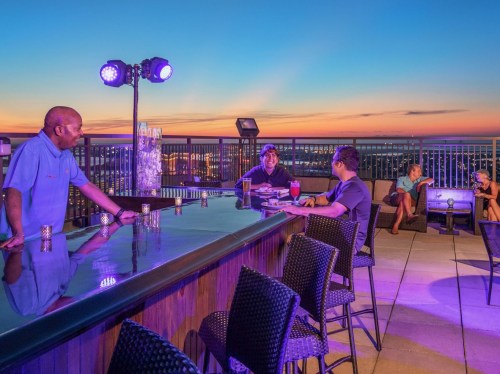 A rooftop bar at sunset with patrons enjoying drinks and a bartender, illuminated by purple lights.