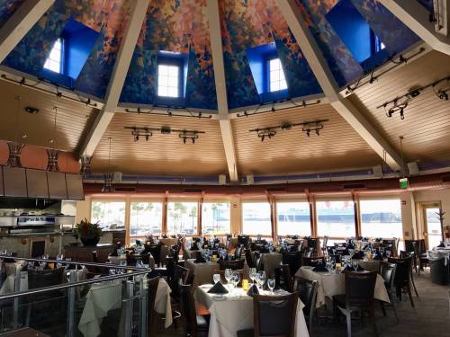Interior of a restaurant with a colorful ceiling, round tables set for dining, and large windows overlooking a waterfront.